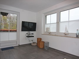 A room with a flat-screen TV mounted on the wall, a window with blinds, a small shelf with decorative items, and a wicker basket on the floor.