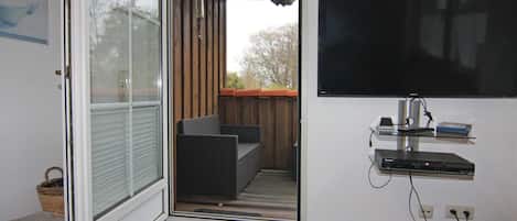 A room with a sliding glass door leading to a balcony, a flat-screen TV mounted on the wall, and a small shelf with electronic devices.