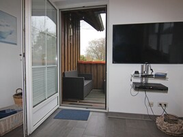 A room with a sliding glass door leading to a balcony, a flat-screen TV mounted on the wall, and a small shelf with electronic devices.