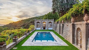 Ein Pool mit einem steinernen Weg, der zu einem Gebäude mit Bogenfenstern und einem Garten mit Weinreben führt.