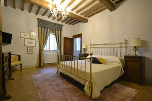 A bedroom with a four-poster bed, wooden beams on the ceiling, a chandelier, a window with curtains, a bedside table with a lamp, and framed pictures on the wall. - Belvilla by OYO Villa Torreone (Cortona)