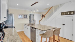 Una cocina con encimera de granito, pileta de acero inoxidable y lavavajillas. Hay taburetes de mimbre y una escalera con un letrero que dice 'BEACH'.