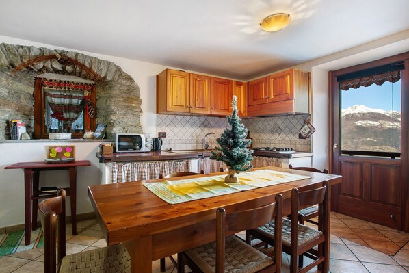 A kitchen with wooden cabinets, a dining table with chairs, a small Christmas tree, and a view of snow-capped mountains through a door.
