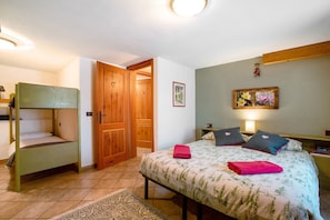 A bedroom with a bunk bed, a single bed with a floral bedspread, a wooden door, a framed picture on the wall, and a bedside table with a lamp.