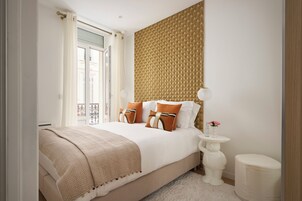 A bedroom with a large bed, a headboard with a geometric pattern, a bedside table with a lamp, and a small white stool.