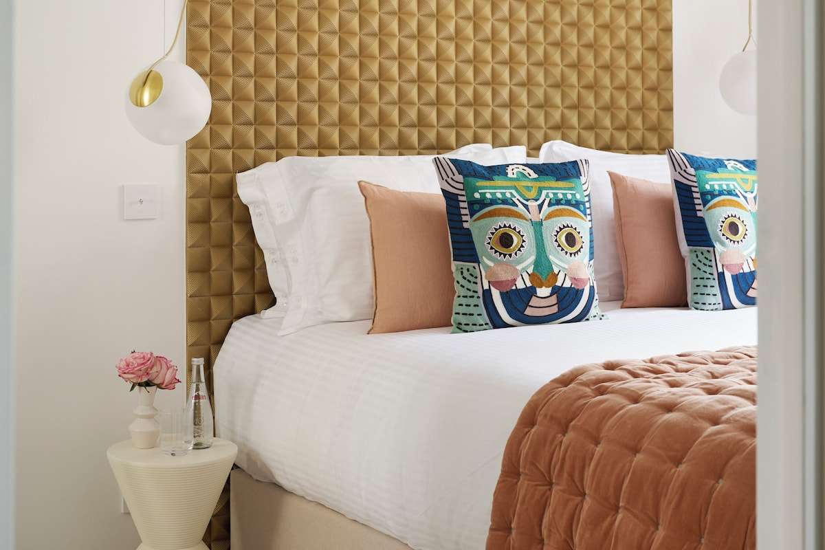 A bedroom with a tufted headboard, a white bedspread, and colorful patterned pillows.