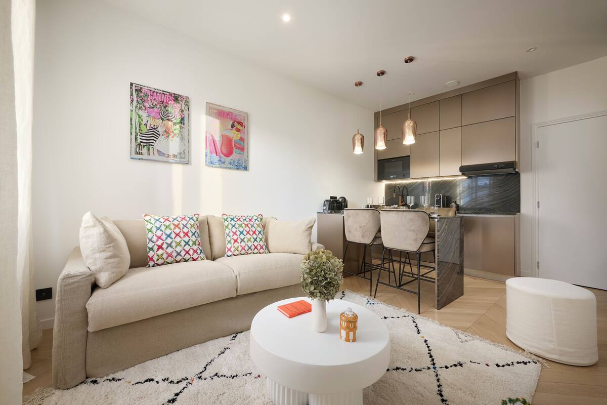 A modern living room with a beige sofa, colorful pillows, a white coffee table with a book and a vase, and a kitchen area with a bar and pendant lights.