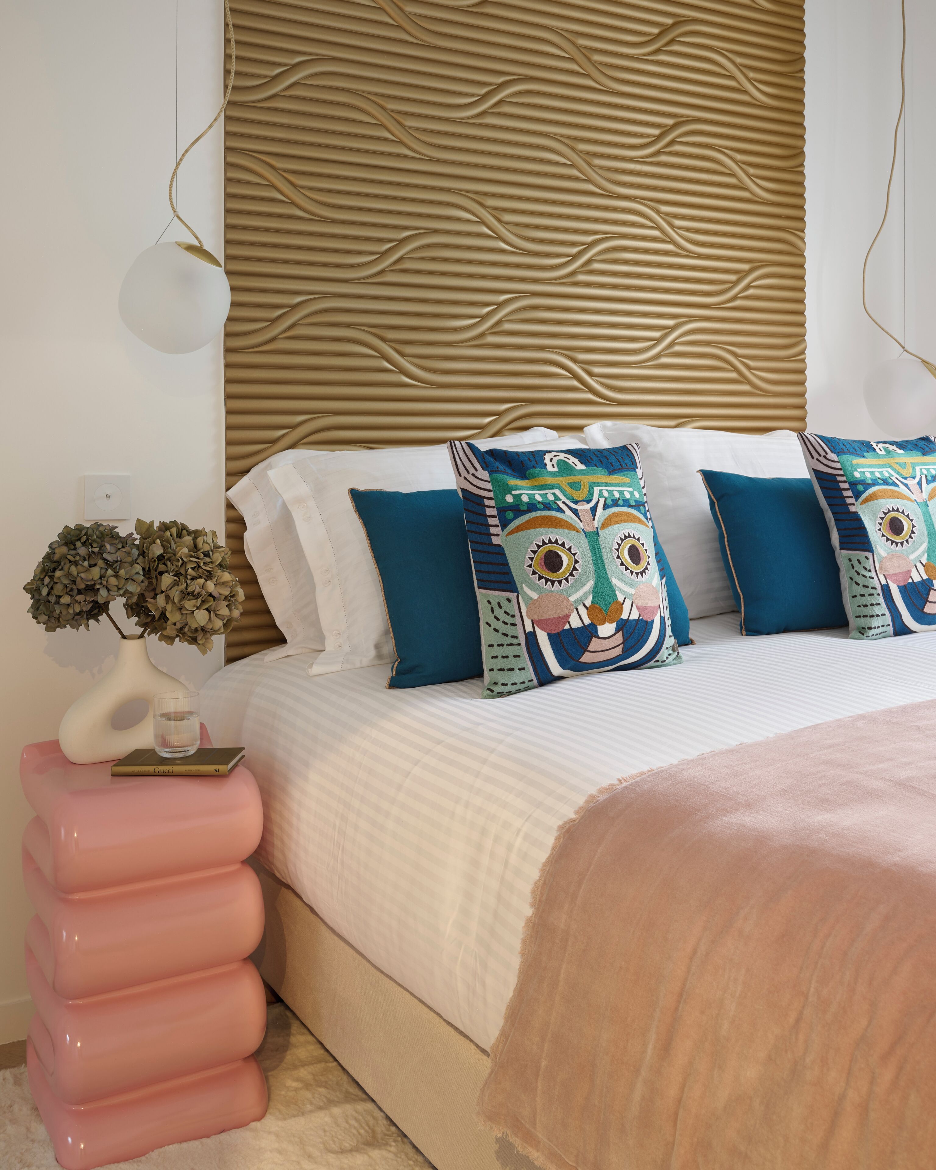 Quarto com cama, almofadas decorativas, mesa de cabeceira com um vaso e parede com textura.
