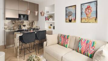 Una cocina moderna con lámparas colgantes de cobre, una zona de comedor con una silla negra y un sofá beige con almohadones coloridos. Hay dos cuadros en la pared.