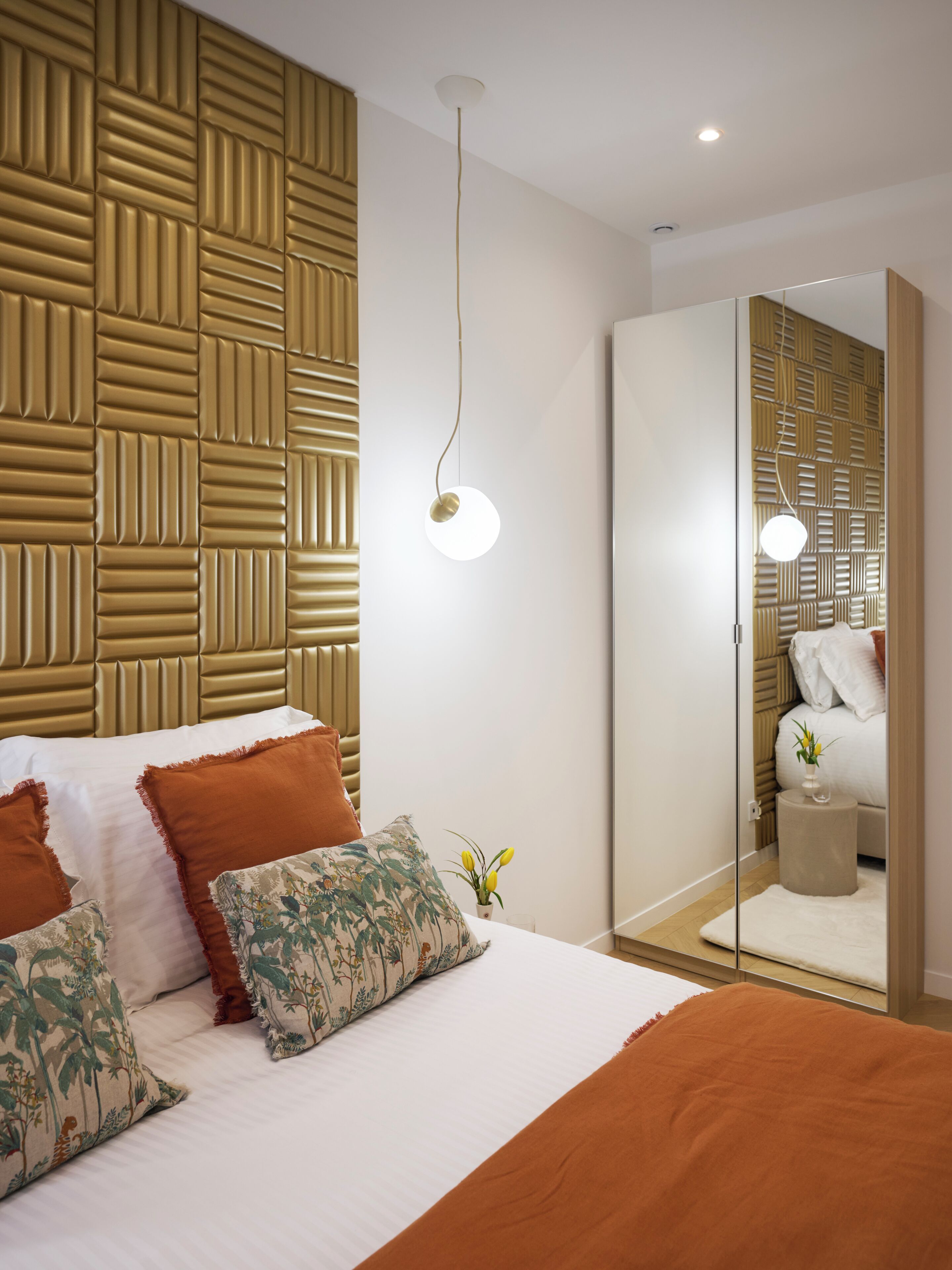 Un dormitorio moderno con un cabecero estampado, una cama grande con muchas almohadas, una mesita de noche con un florero y un armario con espejo.