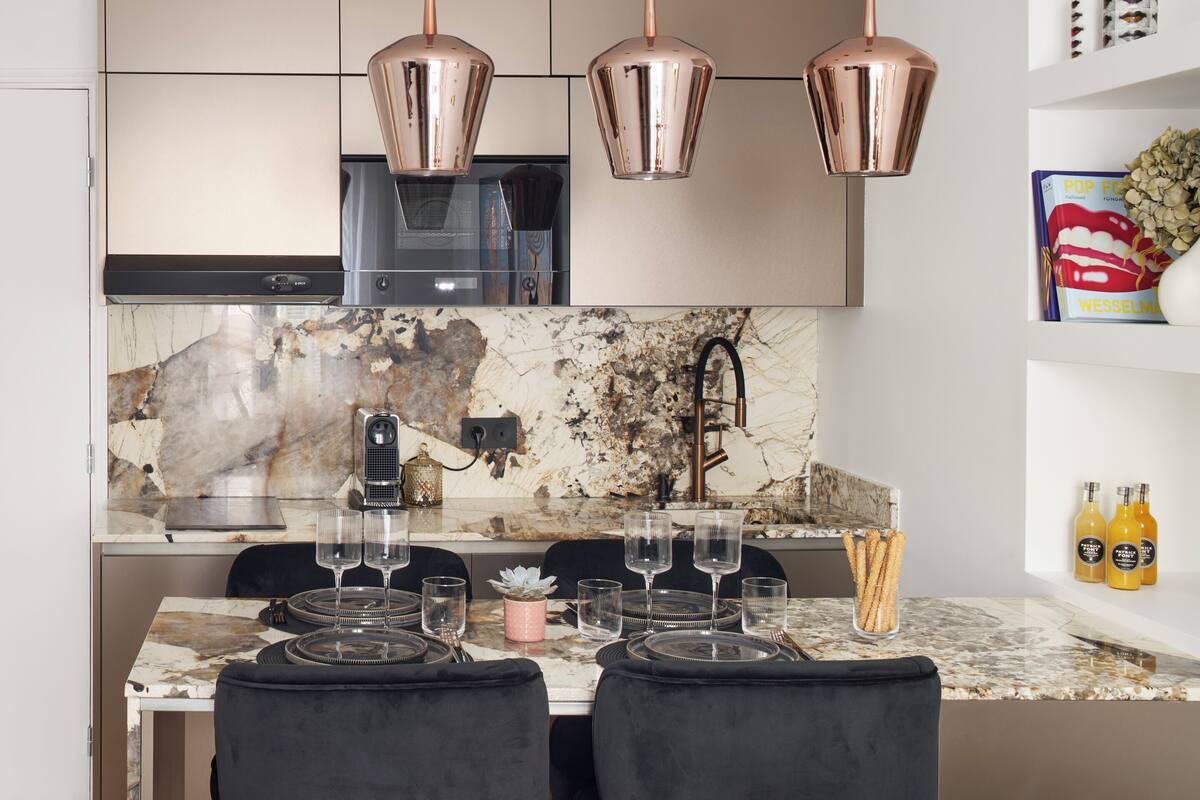 A modern kitchen with a marble backsplash, copper pendant lights, and a dining area with black chairs and a colorful pillow.