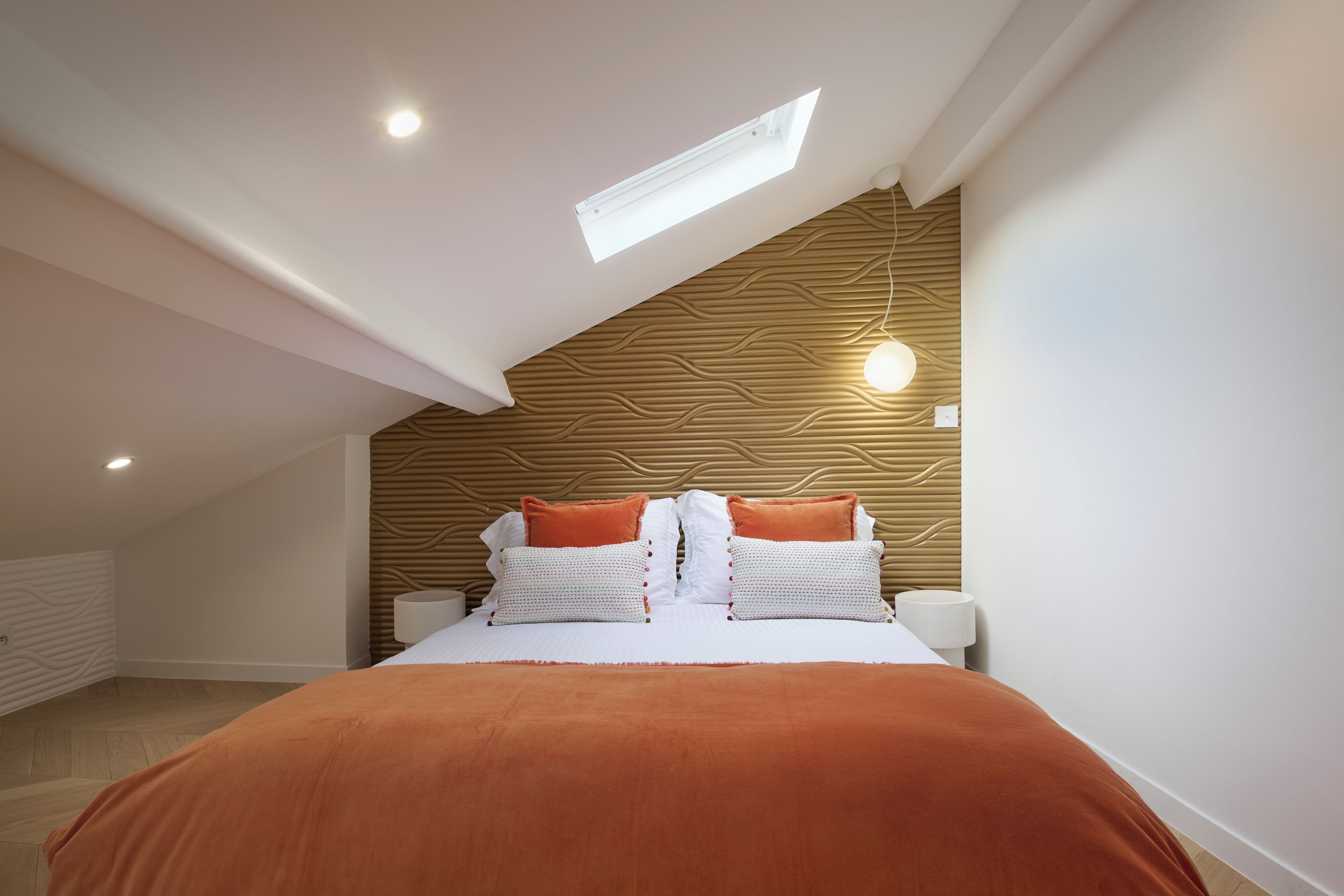 A bedroom with a sloped ceiling, a skylight, a bed with white and orange bedding, and a wooden wall panel.