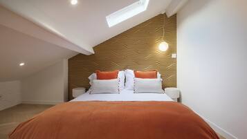 A bedroom with a sloped ceiling, a skylight, a bed with white and orange bedding, and a wooden wall panel.