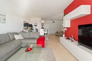 Un salon moderne avec un canapé gris, un plaid rouge, une table basse en verre, un téléviseur à écran plat et un mur d’accent rouge.