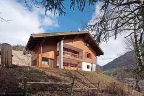 Une maison en bois à deux étages, dotée d’un balcon, située sur une colline avec une chaîne de montagnes en arrière-plan.