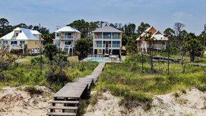 Une propriété située en bord de mer, avec une promenade en bois menant à un espace piscine, le tout entouré de dunes de sable et de végétation.