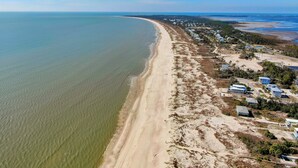 Vue aérienne d’une zone côtière avec une plage de sable, une rangée de maisons et un ciel bleu dégagé.
