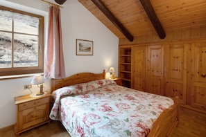 Een slaapkamer met een houten bed, een nachtkastje met een lamp, een raam met uitzicht op een besneeerd landschap en houten inbouwkasten.
