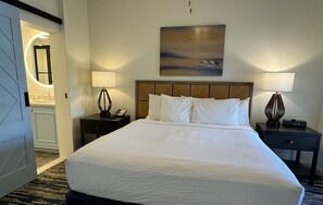 A hotel room with a large bed, two bedside tables with lamps, a painting on the wall, and a bathroom visible through an open door.