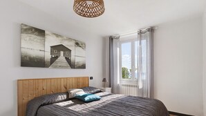 Un dormitorio con cabecera de madera, cama con ropa de cama a rayas, mesita de noche con lámpara, ventana con cortinas y una obra de arte colgada en la pared.