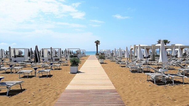 A beach with rows of sun loungers and umbrellas, a wooden walkway, and a palm tree in the distance. - Belvilla by OYO Appartamento Nero (Jesolo)