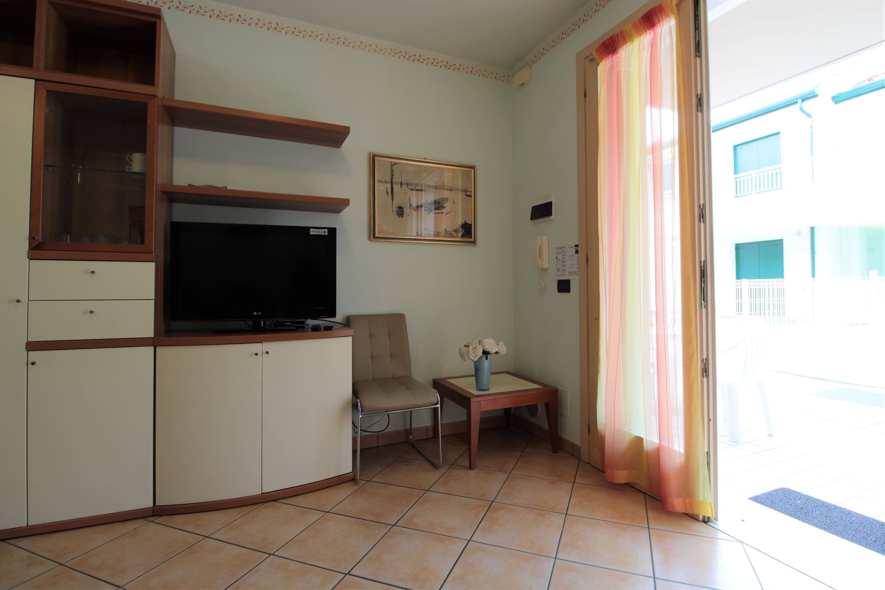 A living room with a TV, a chair, a small table with a vase, and a sliding door to an outdoor area.