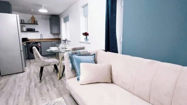 A modern living room with a beige sofa, a white armchair, and a small round table. A kitchen with a washing machine and a dining area with a table and chairs is visible in the background.