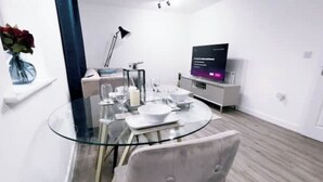 A modern dining area with a glass table, white chairs, and a flat-screen TV on a white stand.