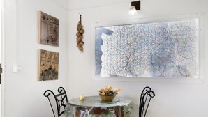 Een eethoek met een tafel, voorzien van een tafelkleed met dessin, twee stoelen, een bloemenmand en wanddecoratie.