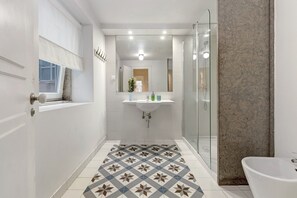 A narrow bathroom with a shower, a sink, and a patterned floor.