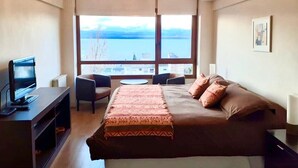 A hotel room with a bed, a desk with a computer, a chair, and a view of a body of water through large windows. - Terrazas del lago I U with a view of Nahuel Huapi Lake (San Carlos de Bariloche)