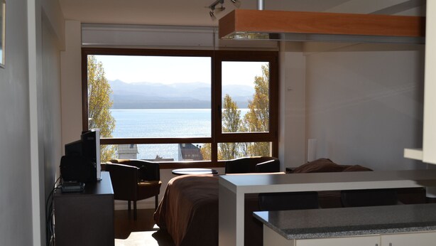 A modern room with a large window, a bed, a desk, and a kitchenette. - Terrazas del lago I U with a view of Nahuel Huapi Lake (San Carlos de Bariloche)