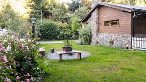 Een rustieke blokhut met stenen fundering en houten buitenkant, omgeven door een verzorgde tuin met vuurkorf en lantaarn.