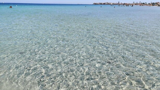 A beach with clear shallow water, a distant shoreline with buildings, and a few people in the water.