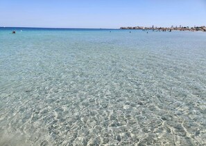 Une plage avec une eau claire et peu profonde, une côte lointaine parsemée de bâtiments et quelques personnes dans l’eau.