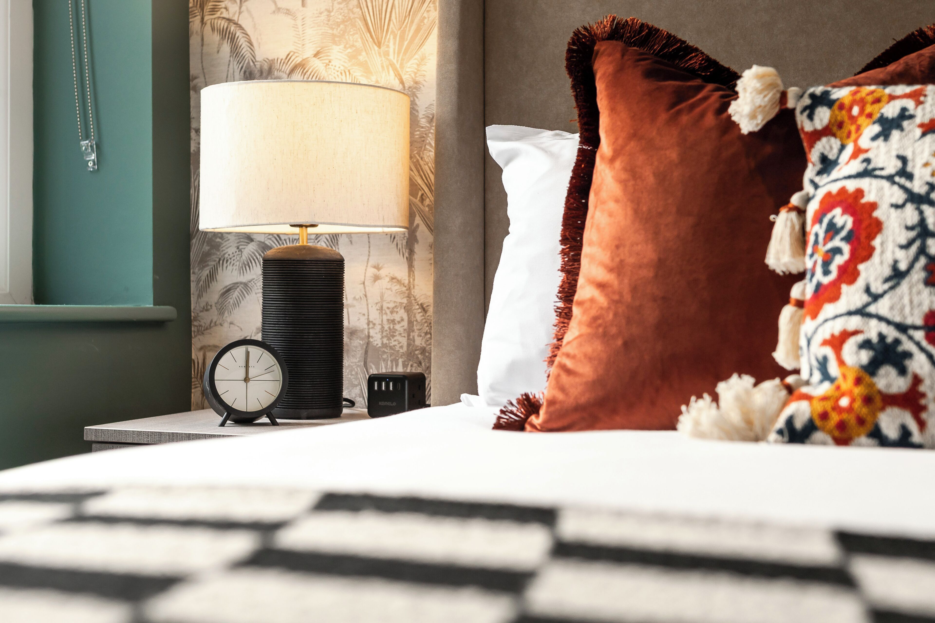 A neatly made bed with a patterned pillow, a bedside lamp, and a black alarm clock on a bedside table.