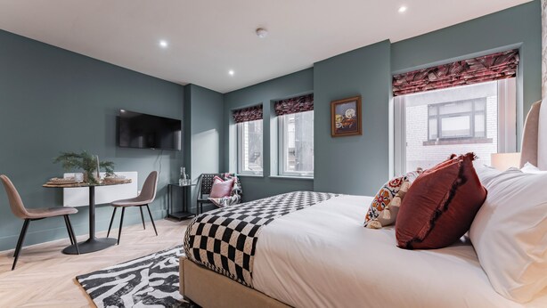Un dormitorio con cama, escritorio, sillas, televisor y ventana con cortinas.