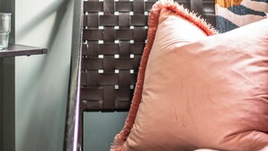 Una silla de mimbre con un cojín rosa y una manta colorida, colocada frente a una pared verde claro.