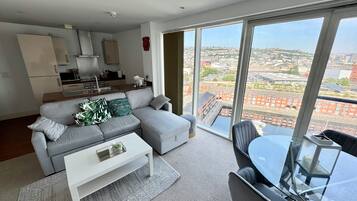 Una sala de estar moderna con un sofá gris, una mesa de centro blanca y una mesa de comedor de vidrio. Se ve una cocina al fondo, con vistas a un paisaje urbano a través de amplios ventanales.