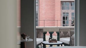 Et værelse med en seng, et lille bord og to stole. Der er en balkon med udsigt til en murstensbygning og en hængende lampe.