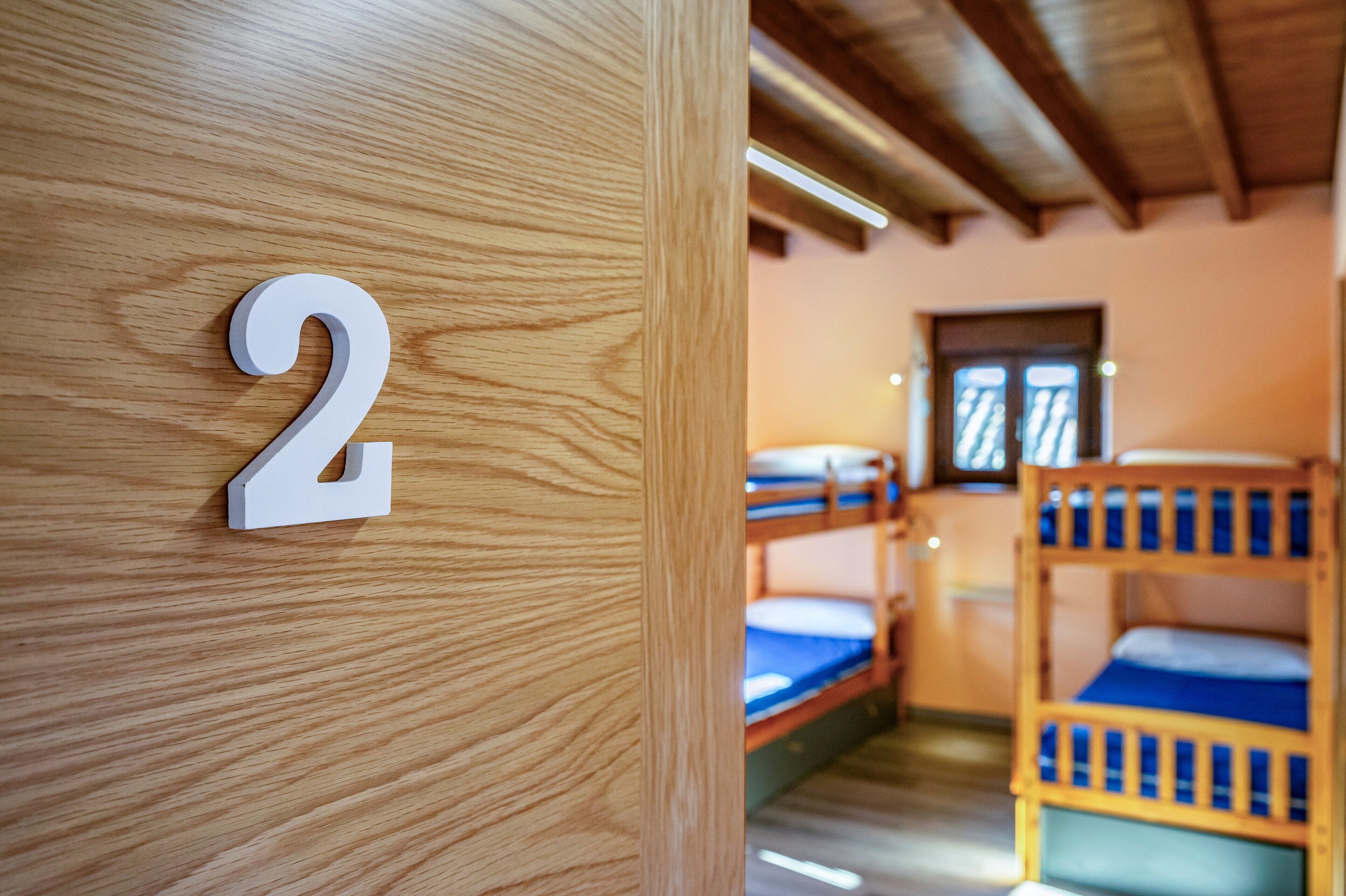 A wooden door with a number '2' indicating a room number, bunk beds with blue bedding, and a window with blinds.