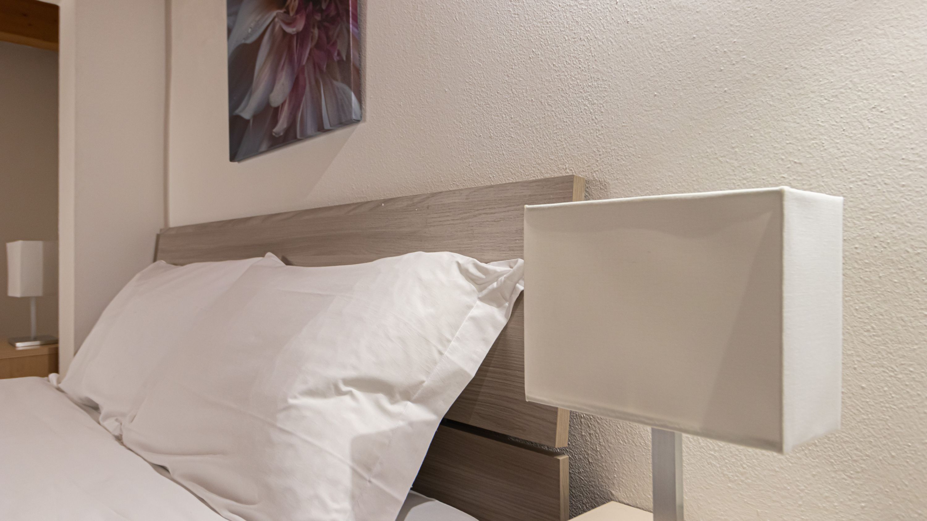 A neatly made bed with white linens and a wooden headboard. A white bedside table with a lamp is visible to the left.