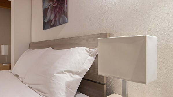 En pænt redt seng med hvide sengetøj og en trægavl. Et hvidt natbord med en lampe er synligt til venstre.