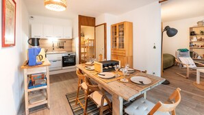 Une cuisine compacte dotée d’appareils électroménagers modernes, un coin repas avec une table et des chaises en bois, et un coin salon confortable avec un fauteuil à bascule.