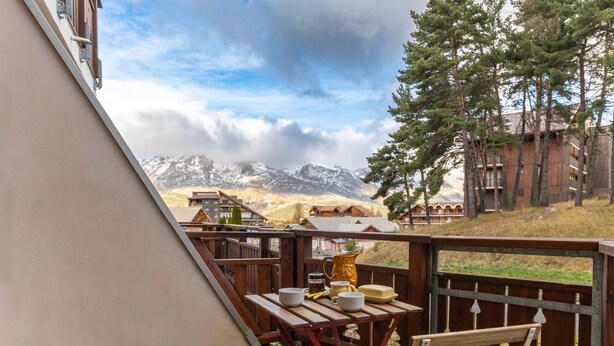 Une terrasse en bois avec une table dressée pour le repas, offrant une vue sur un paysage montagneux avec des bâtiments en arrière-plan.