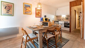 Un coin repas avec une table en bois, quatre chaises et un tapis à rayures. Deux affiches encadrées ornent le mur, ainsi qu’un luminaire suspendu. On aperçoit également une cuisine au fond.