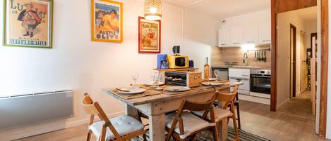 Un coin repas avec une table en bois, quatre chaises et un tapis à rayures. Deux affiches encadrées ornent le mur, ainsi qu’un luminaire suspendu. On aperçoit également une cuisine au fond.
