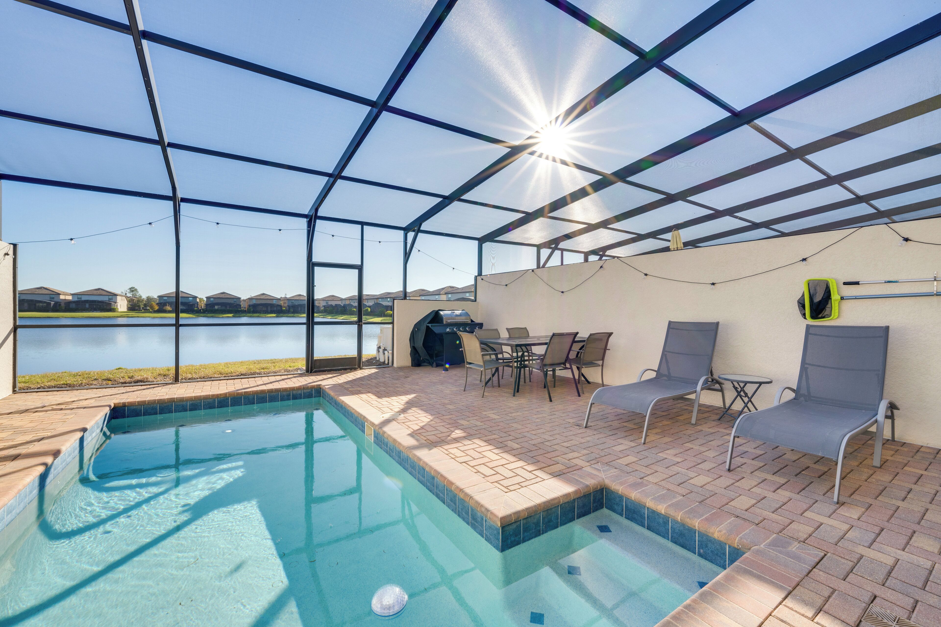 A covered outdoor swimming pool with a view of a lake, surrounded by patio furniture and a grill.