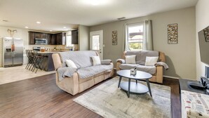 Ein modernes Wohnzimmer mit einem beigefarbenen Ecksofa, einem runden Couchtisch und einer Küchenzeile mit dunkelholzbefischten Schränken und Edelstahlgeräten.