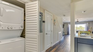 A laundry room with a front-loading washer and dryer, a white door, and a hallway leading to a kitchen area with a window. - Walk to Okaloosa Island Beach: Coastal Condo! (Fort Walton Beach)